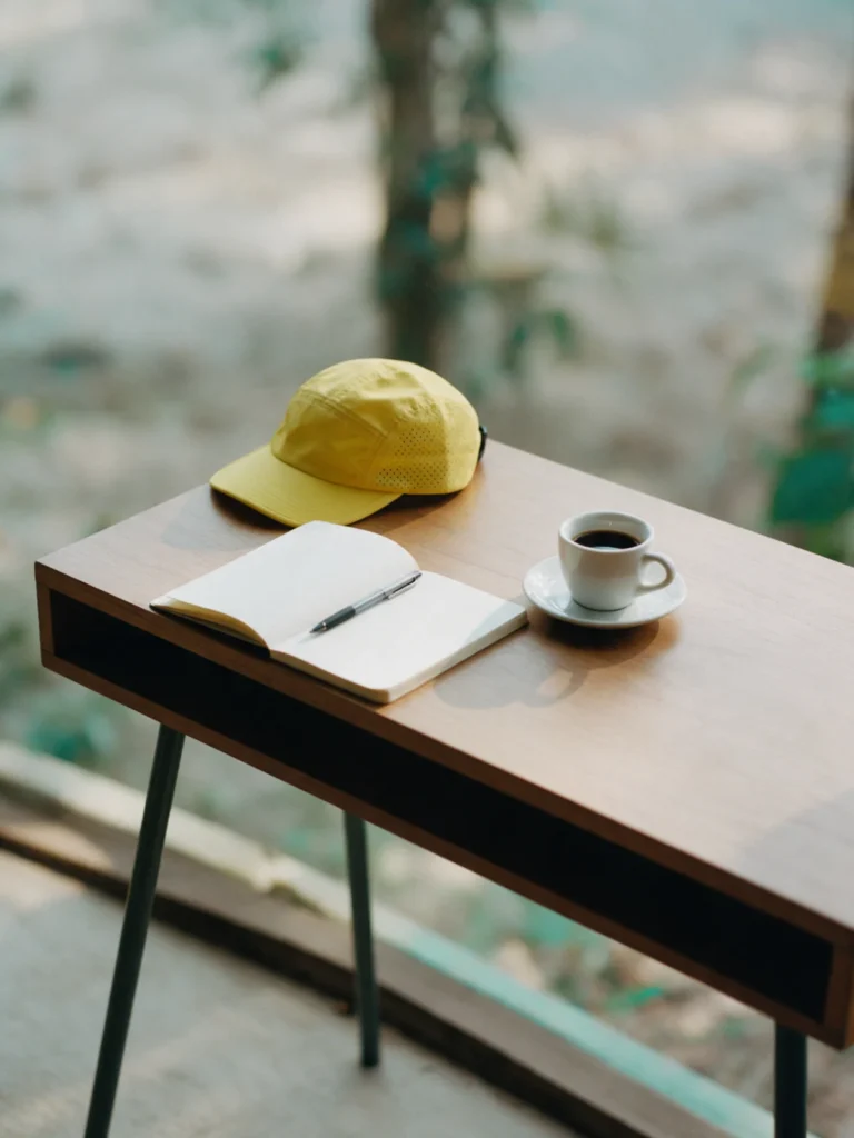 Mesa de madeira com caderno aberto, caneta, café e um boné amarelo